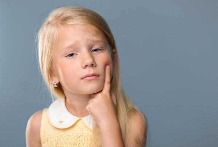 A child with blonde hair wearing a yellow dress touches their cheek with one finger, looking pensive against a gray background.