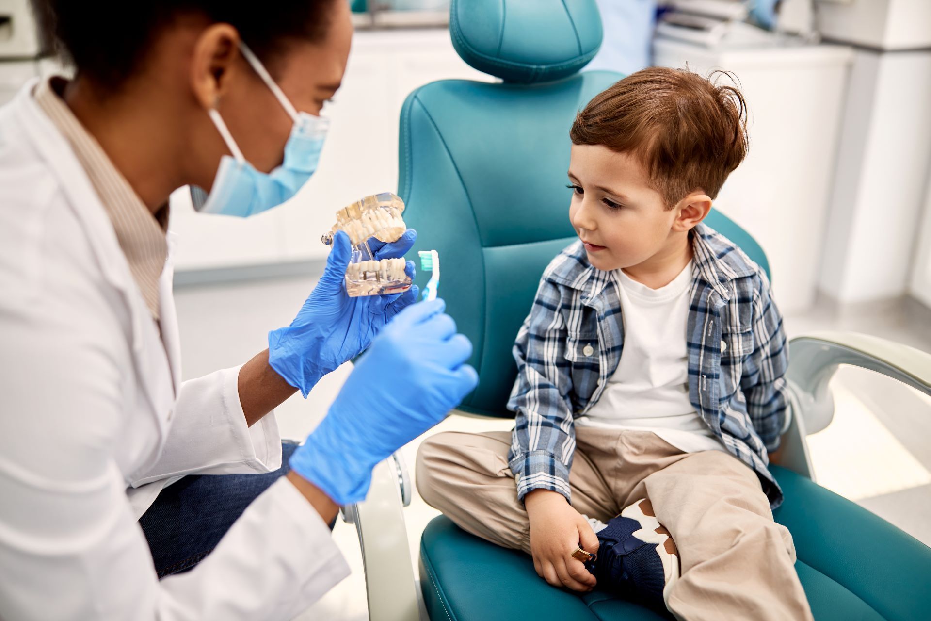 A dental professional in a white coat and blue gloves shows a child how to brush teeth using a dental model.