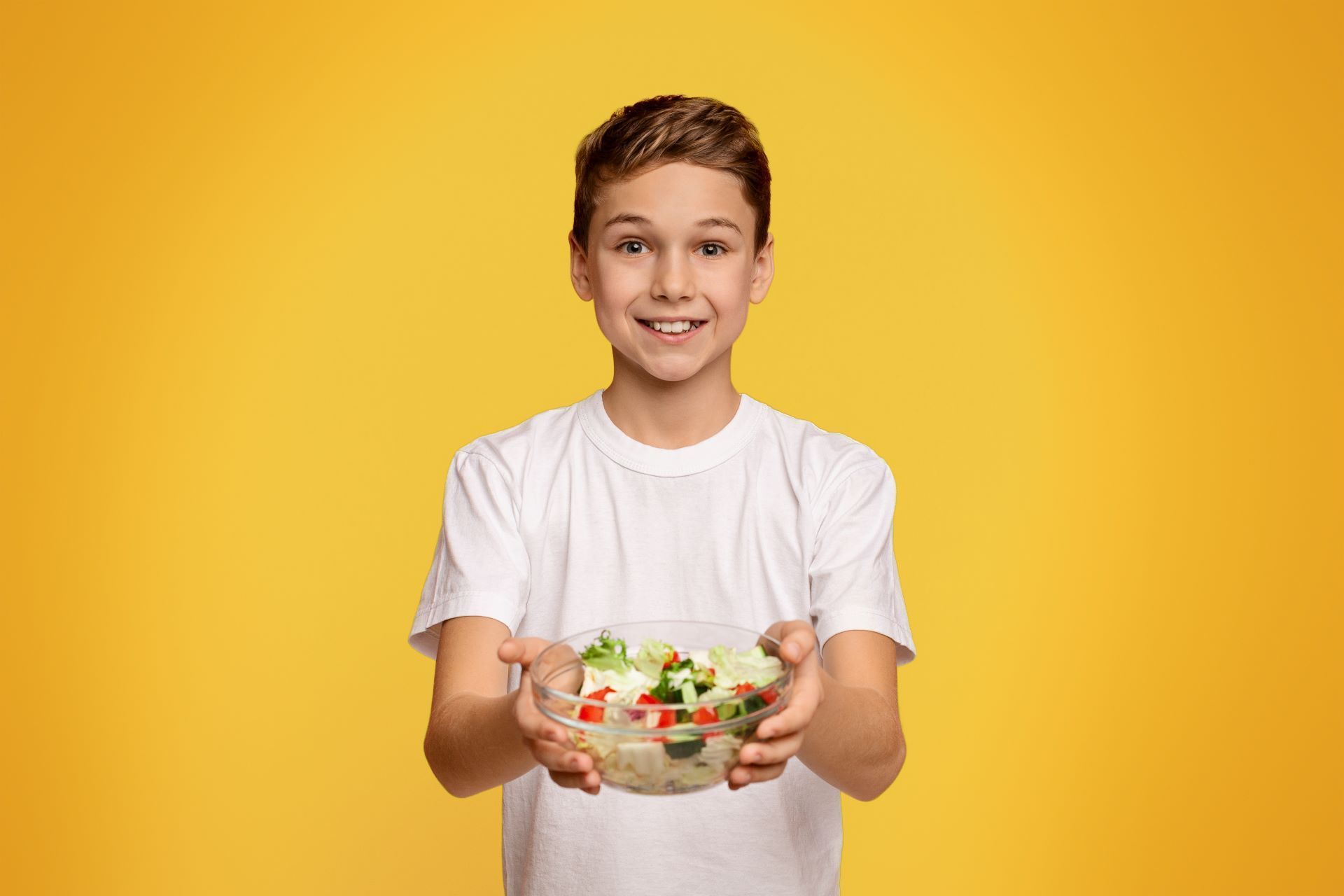 A person in a white t-shirt smiles while holding out a bowl of fresh salad against a bright yellow background.