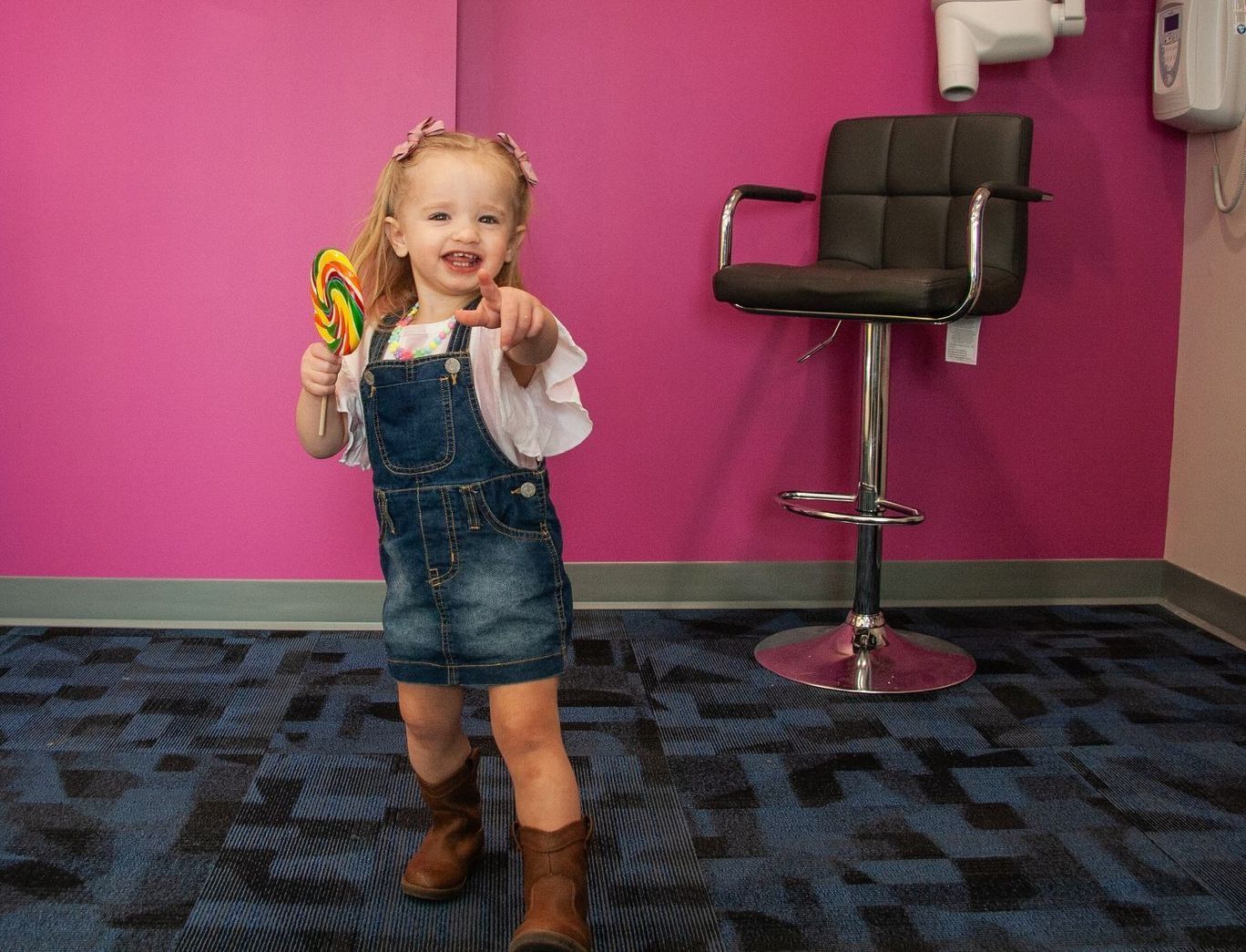 A toddler in denim overalls holds a lollipop and points while standing in front of a bright pink wall and a bar stool.