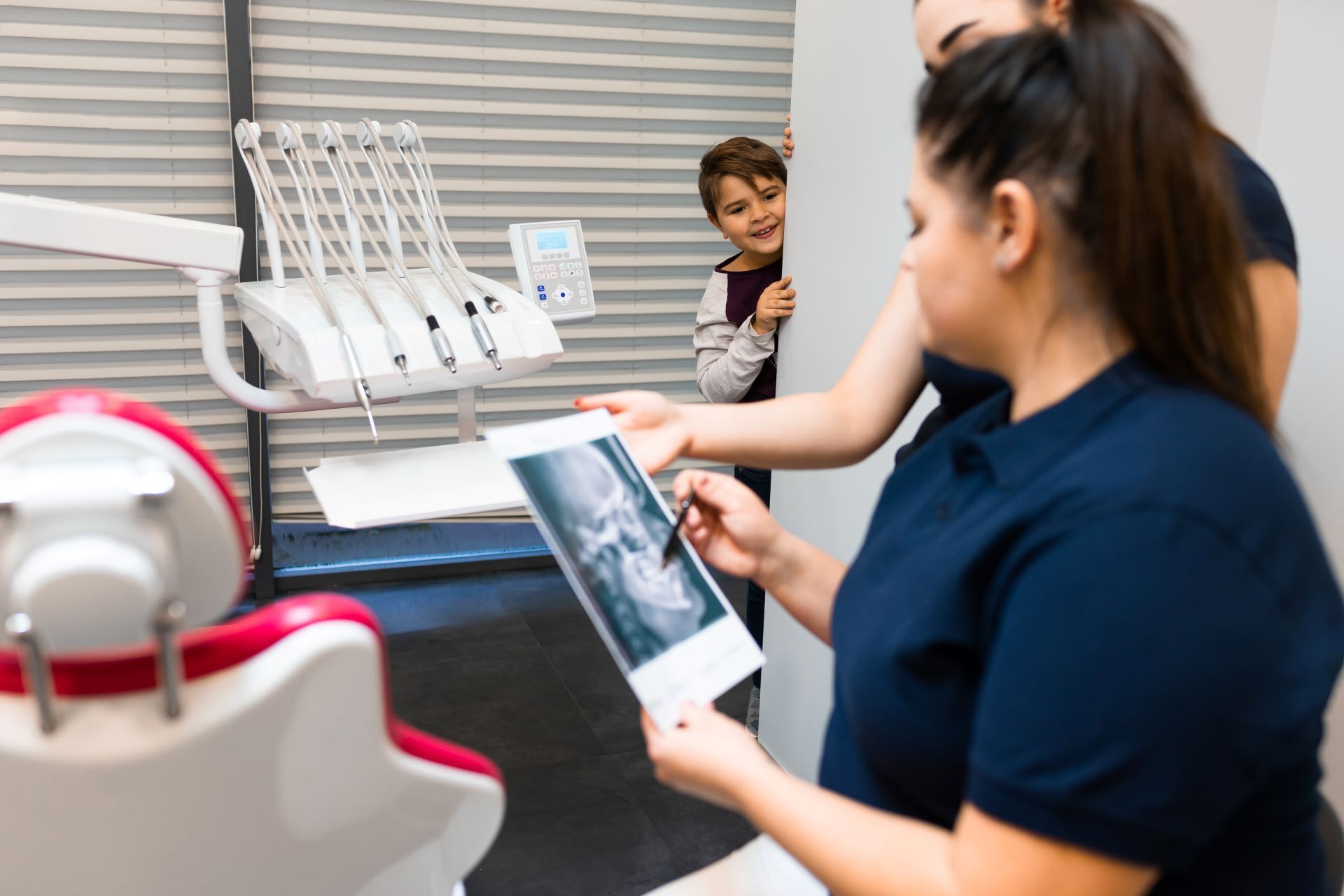 A dental professional holds an X-ray image for consultation while a child peers from behind a wall in a dental office.