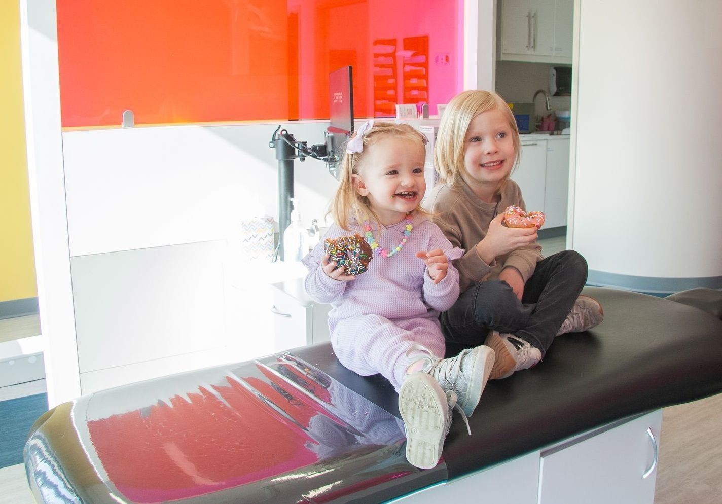 Two children sit on a medical exam table in a bright room, smiling while holding small treats.