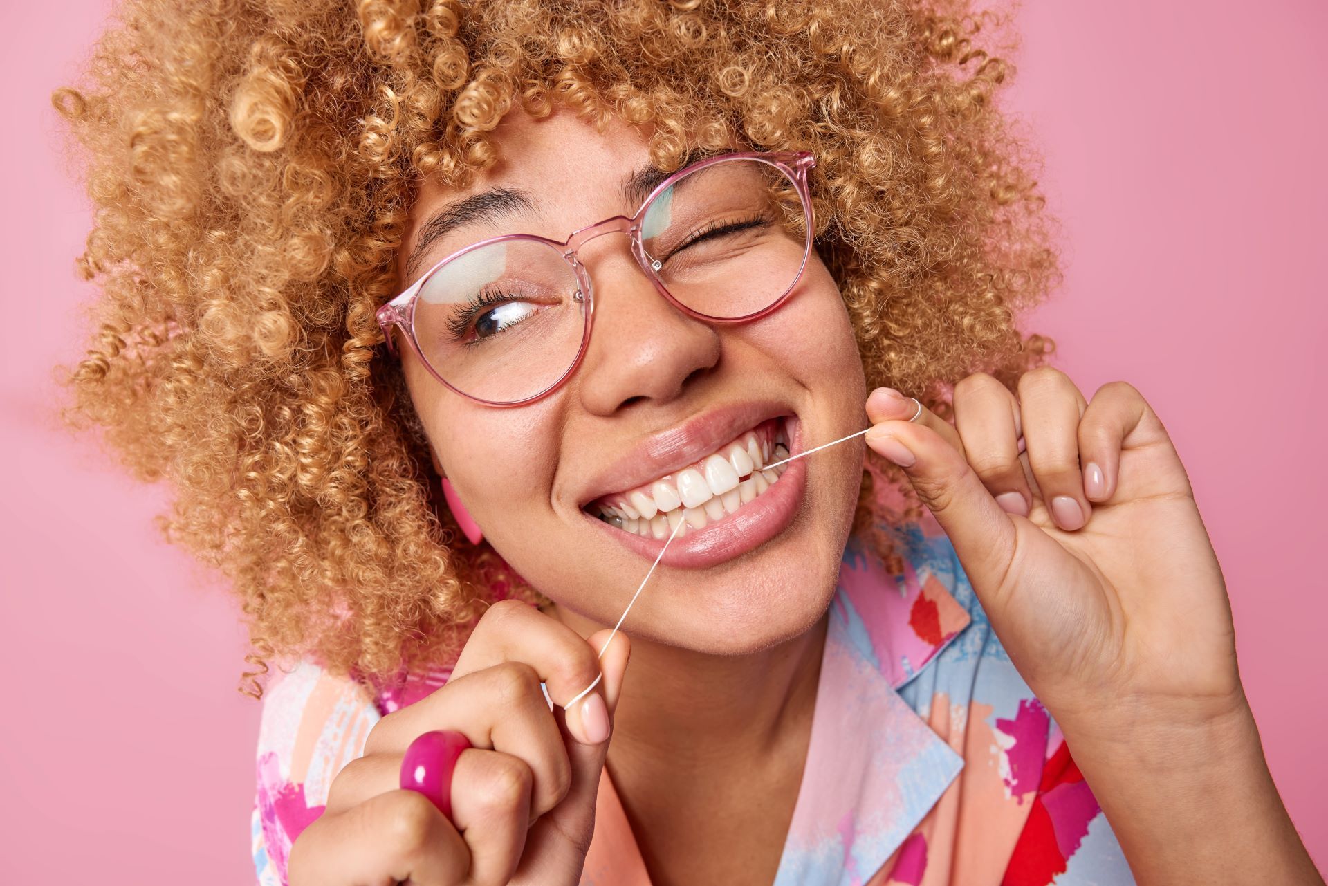 A person with curly hair wearing glasses winks while flossing their teeth against a plain pink background.