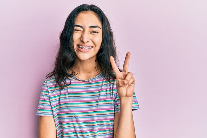 A person with braces winks and holds up a peace sign against a solid light purple background.