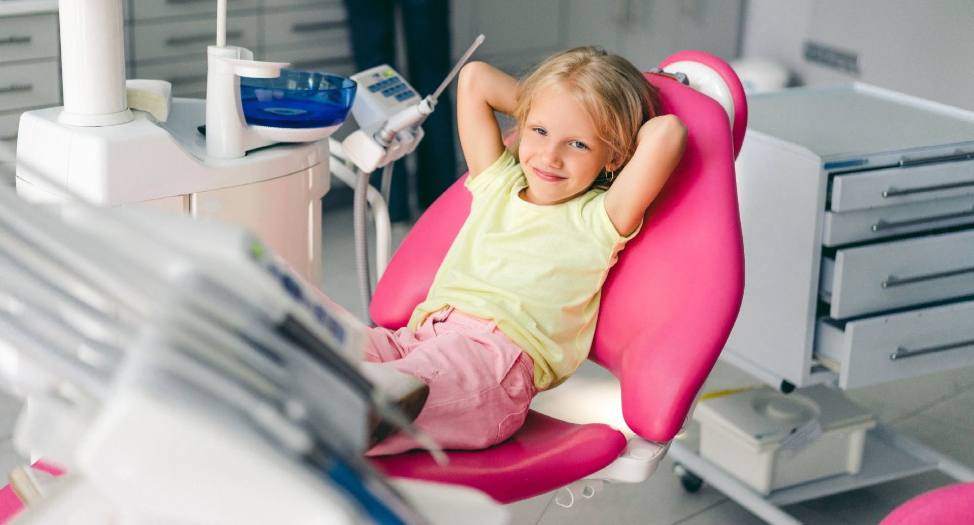 A smiling child sitting with hands behind their head in a pink dental chair inside a bright office.