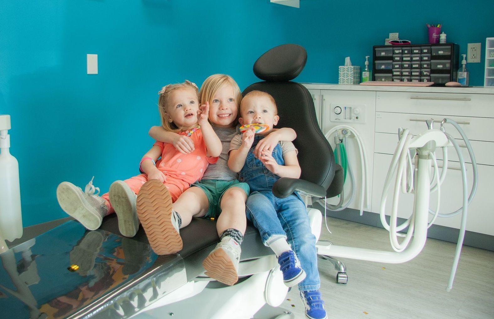 Three children sit together on a black dental chair in a room with bright teal walls and white dental equipment.