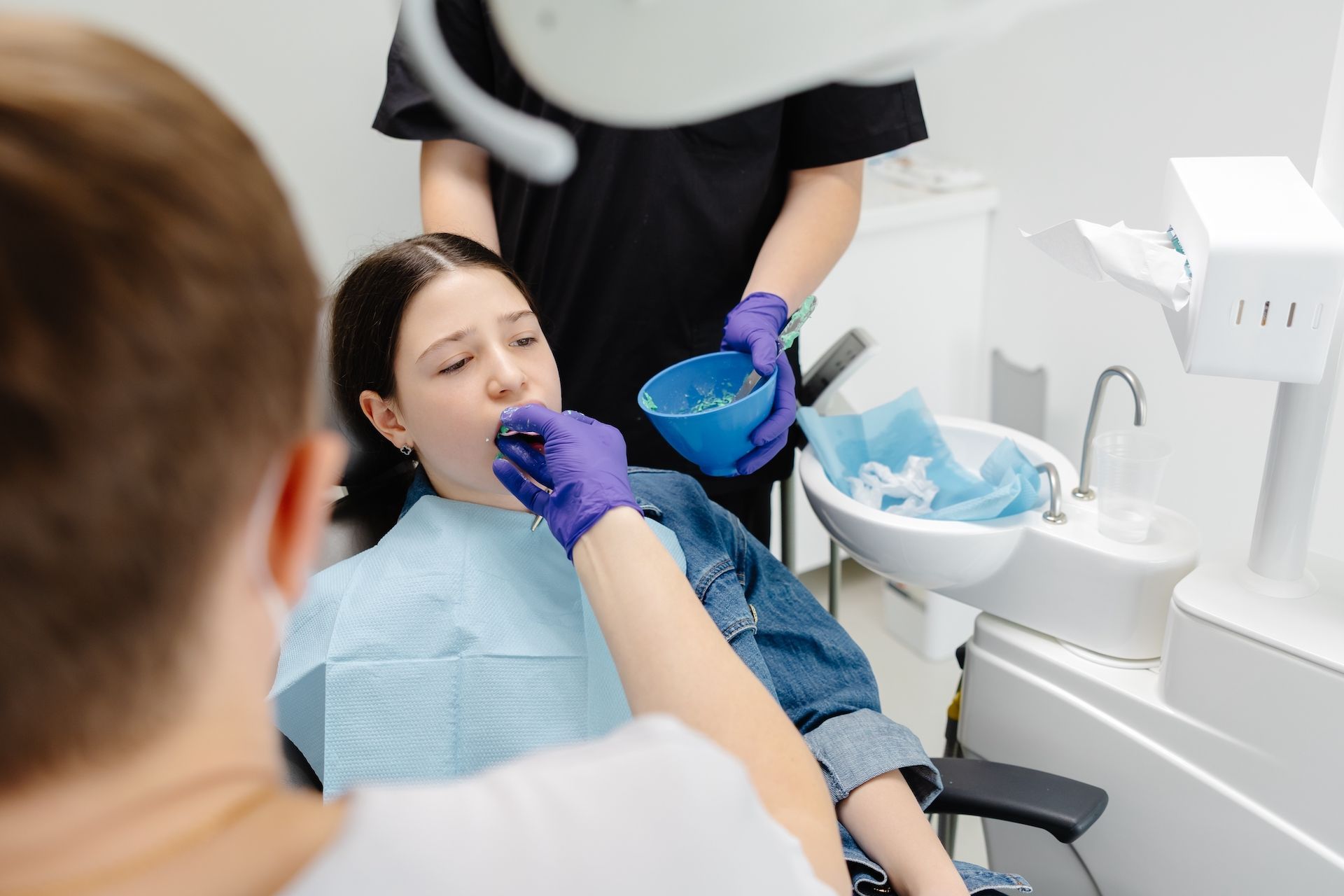 Dental professional applying treatment to a patient's teeth in a clinical setting, both wearing purple gloves.