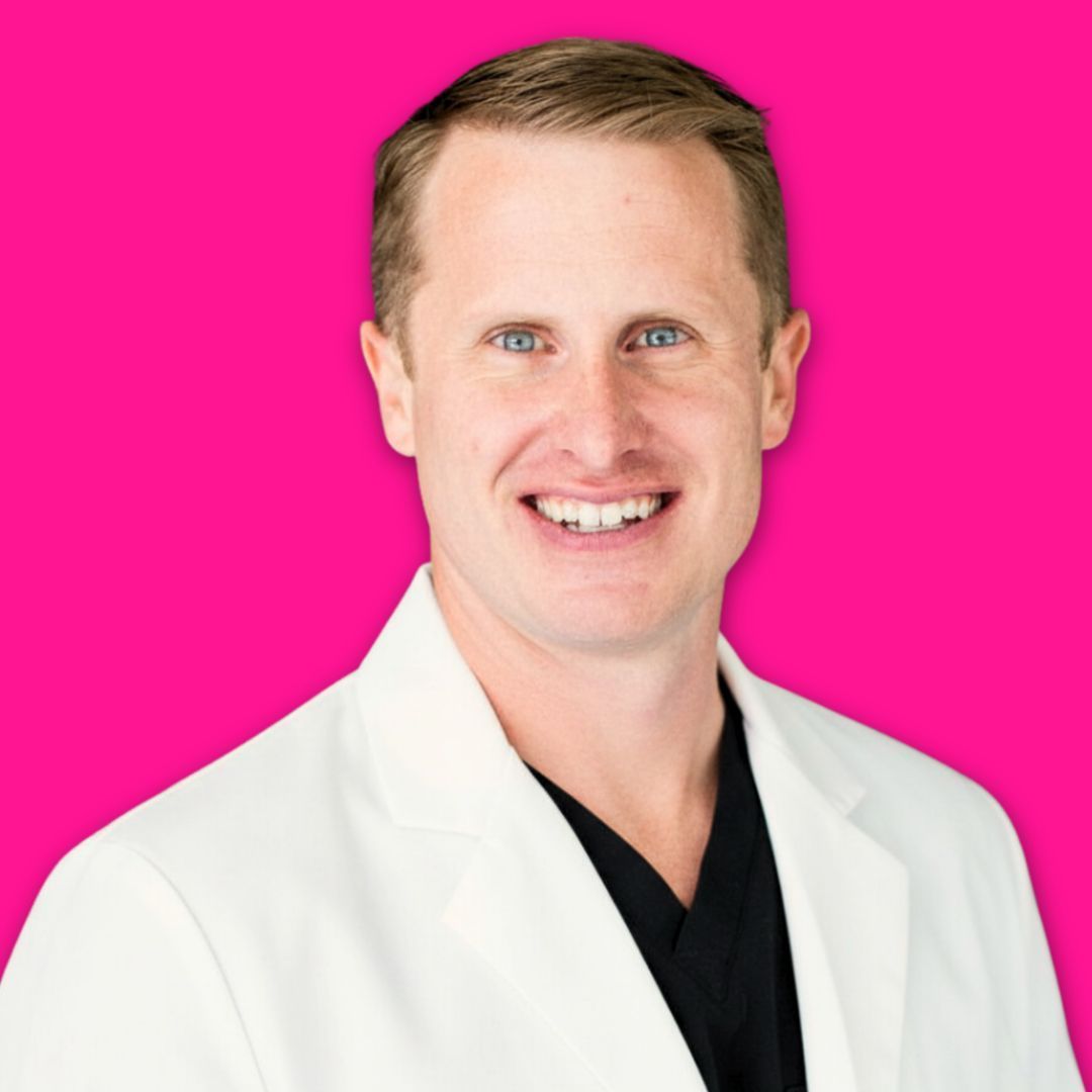 A smiling person wearing a white lab coat over a black shirt against a solid bright pink background.