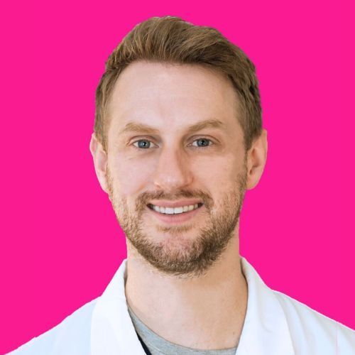 A smiling person with short light hair and a beard, wearing a white lab coat against a bright pink background.