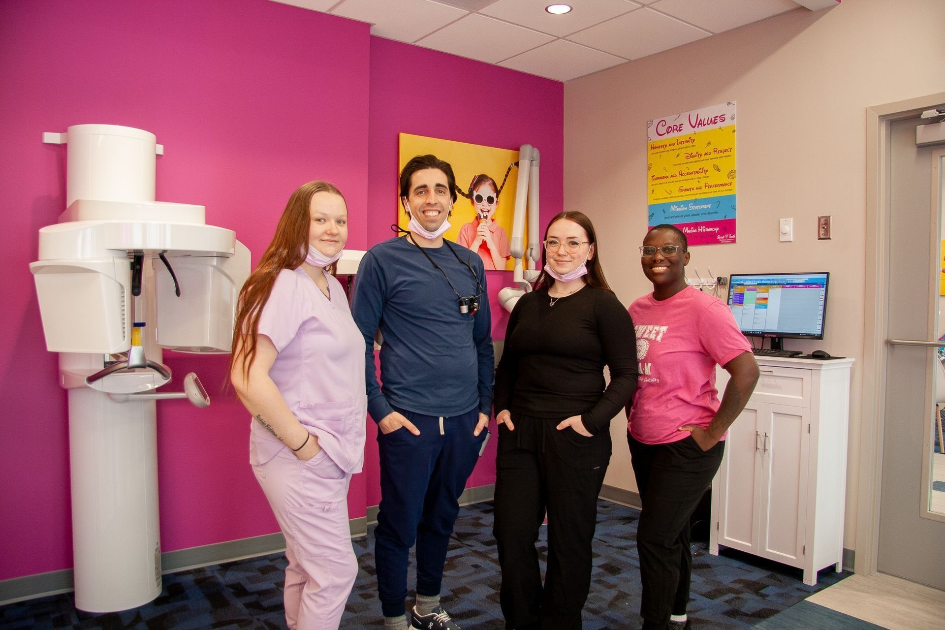 Four dental office staff smiling in a room with a bright pink wall and a panoramic X-ray machine.
