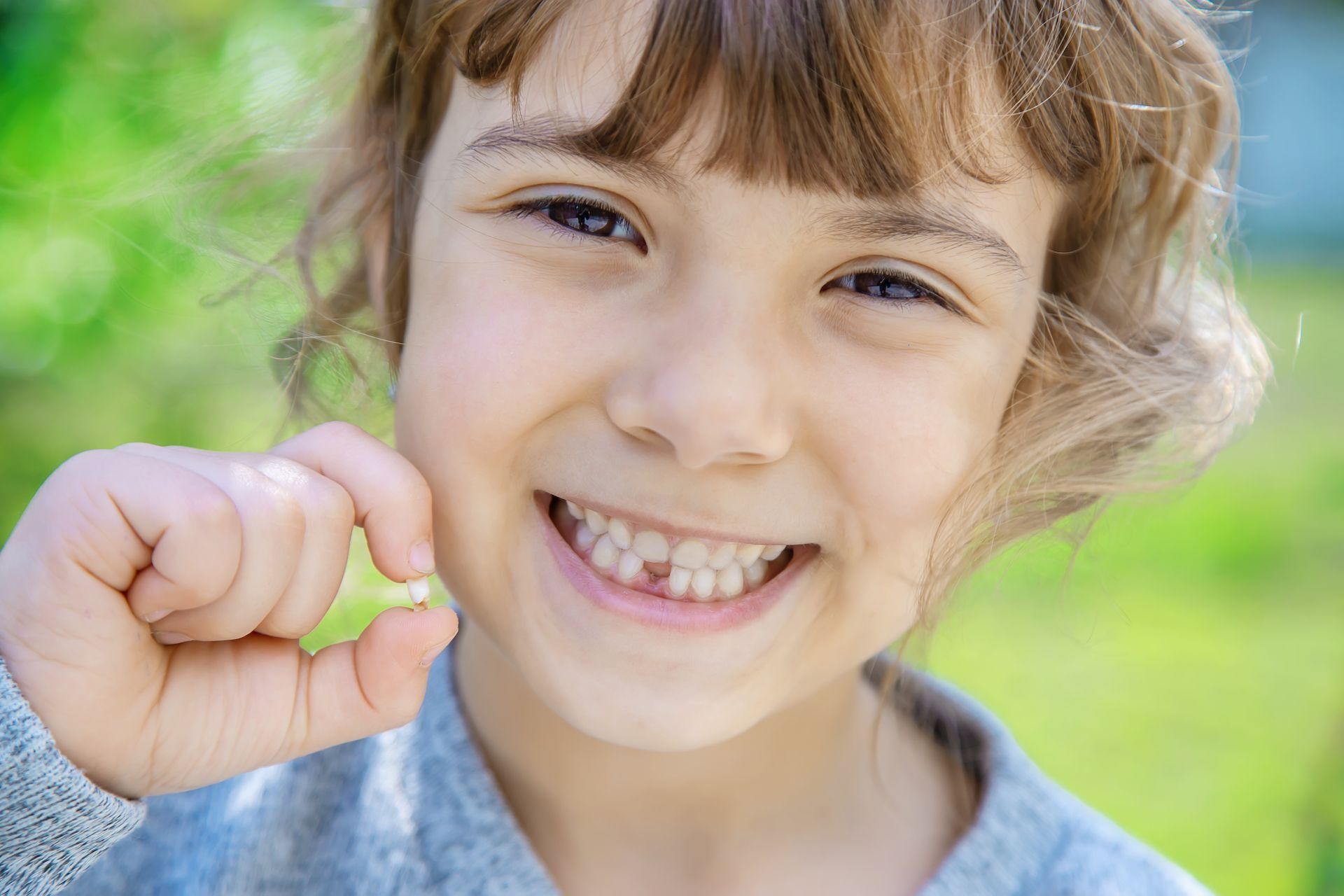A smiling person holding a lost tooth between their fingers with a blurred green outdoor background.