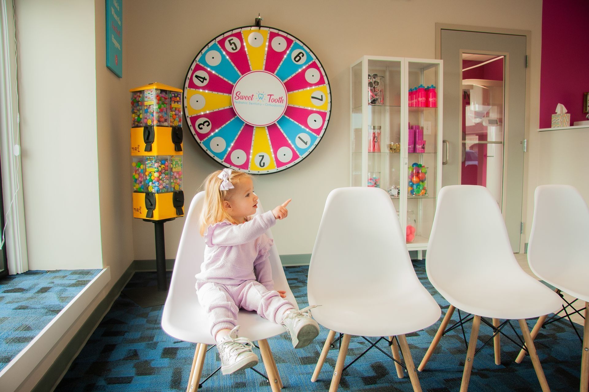 A child points at a colorful prize wheel in a waiting room with chairs and a gumball machine.