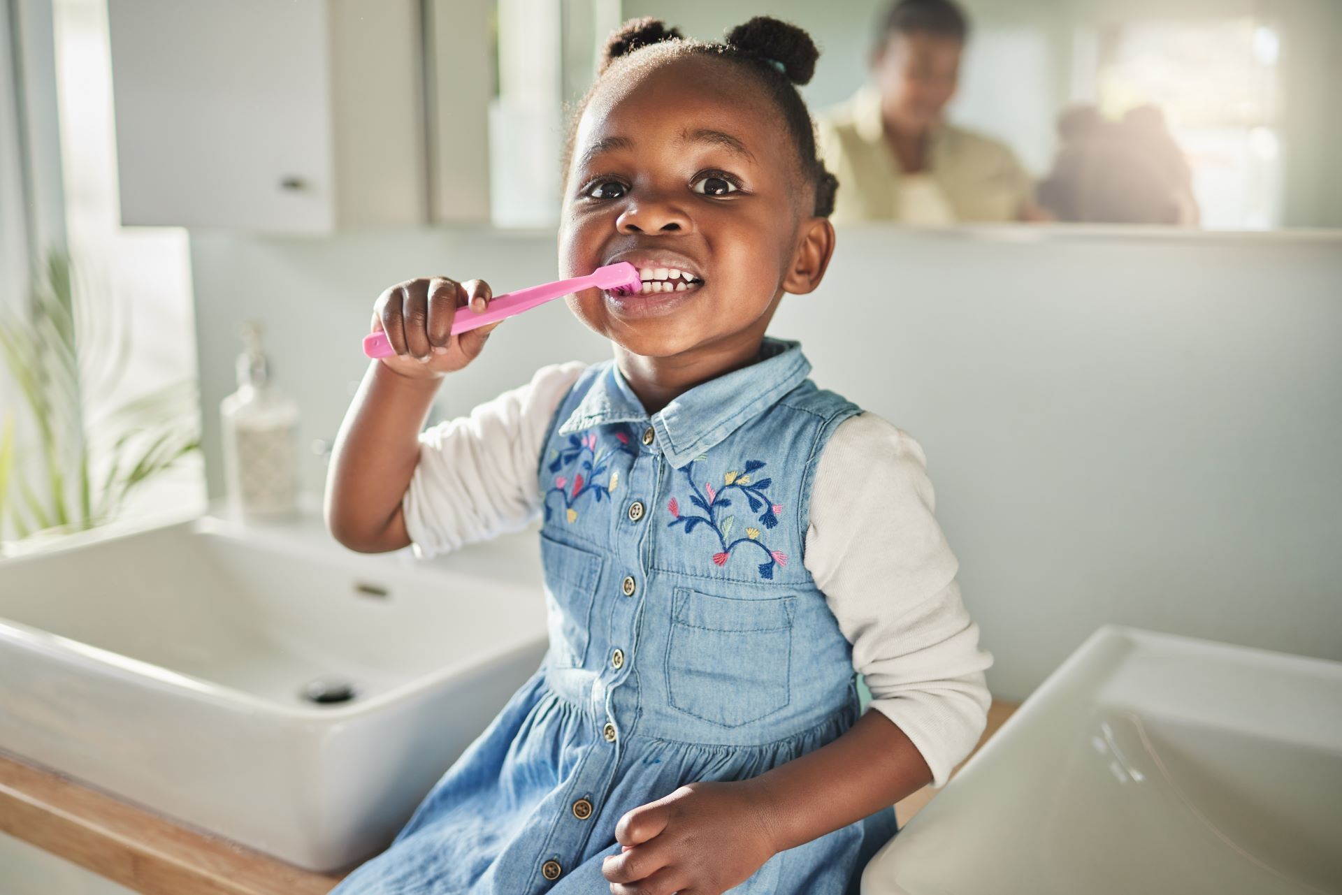A young person in a denim dress smiles while brushing their teeth with a pink toothbrush in a bathroom.