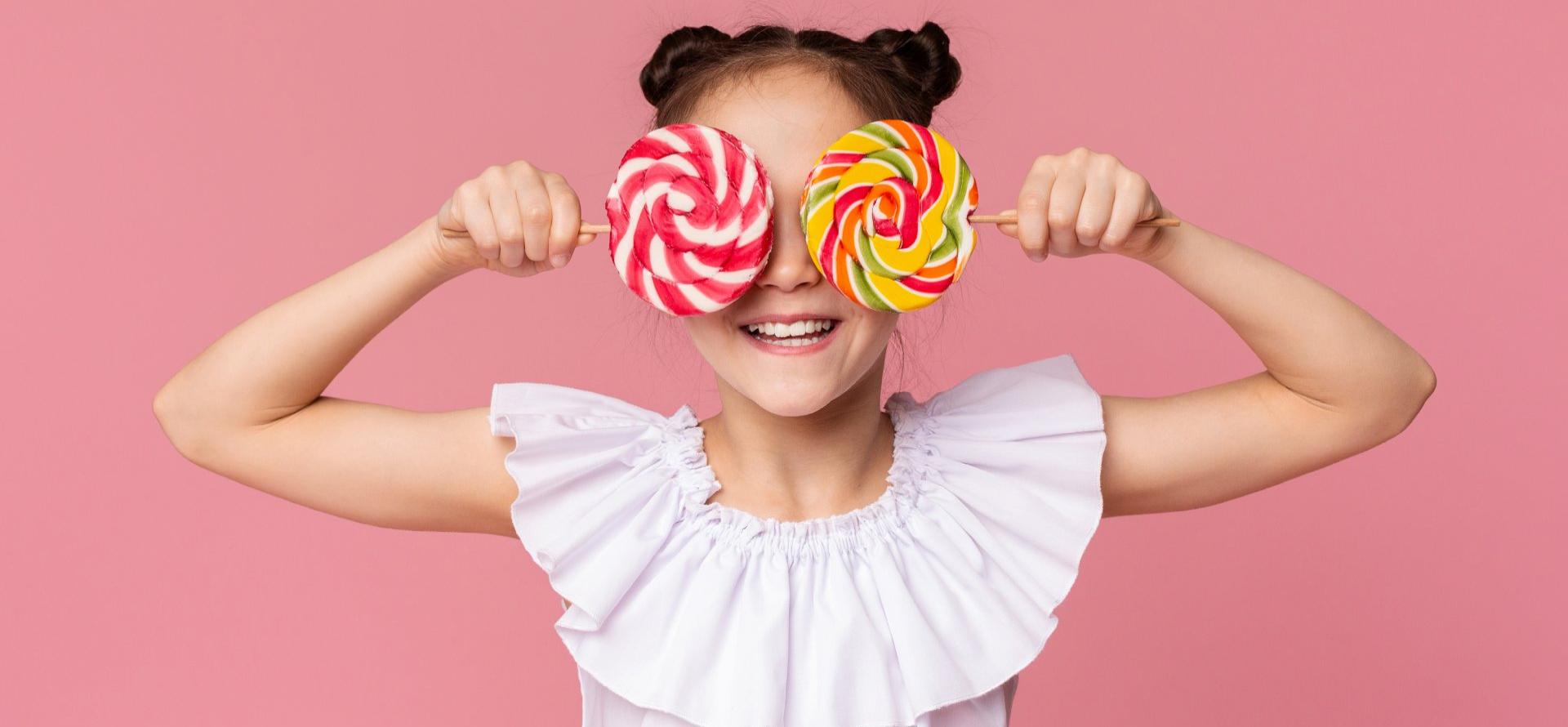 A smiling person holds a pink-and-white swirl lollipop over one eye and a multi-colored swirl lollipop over the other.