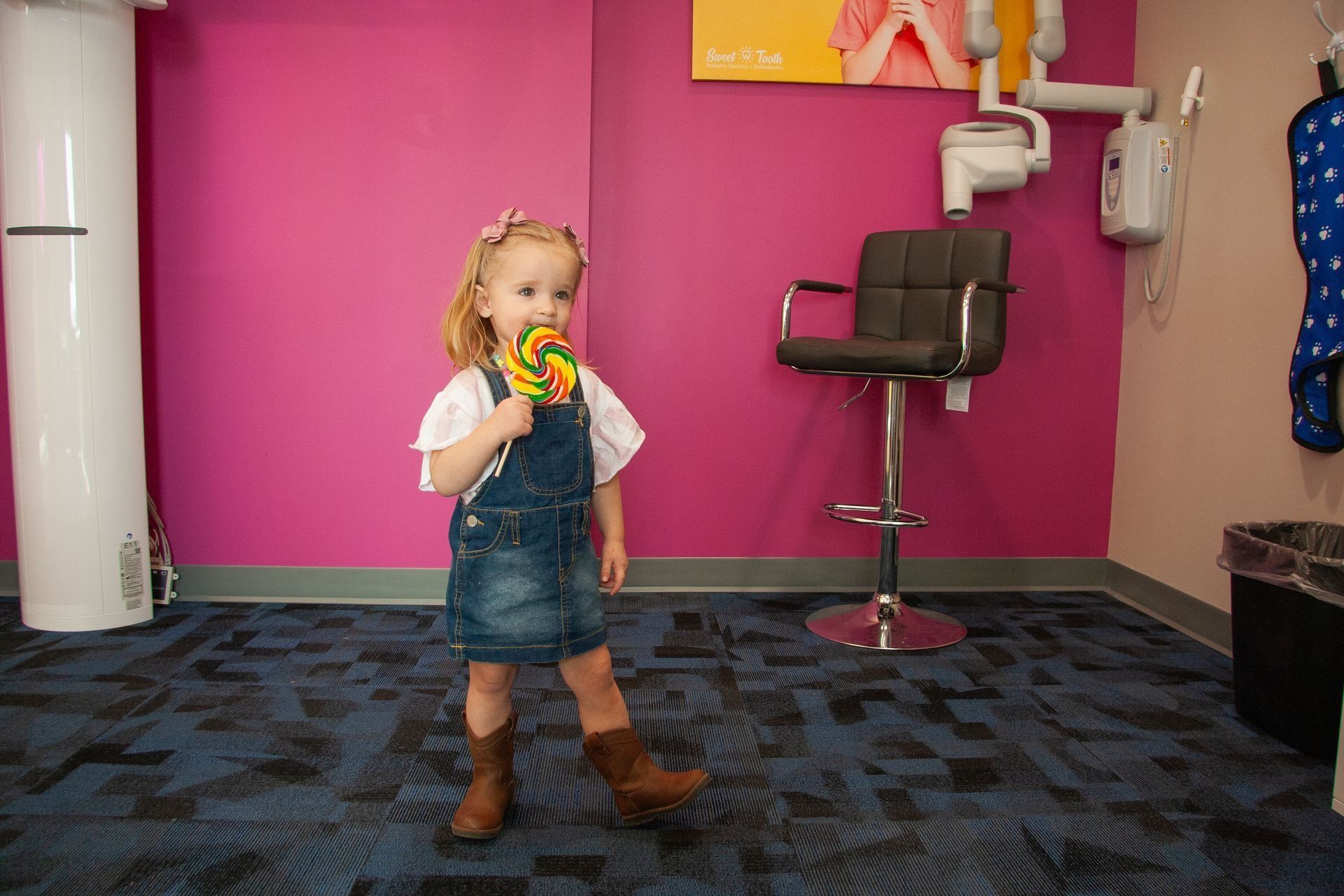 A young person wearing denim overalls and brown boots holding a lollipop in a dental office with hot pink walls.