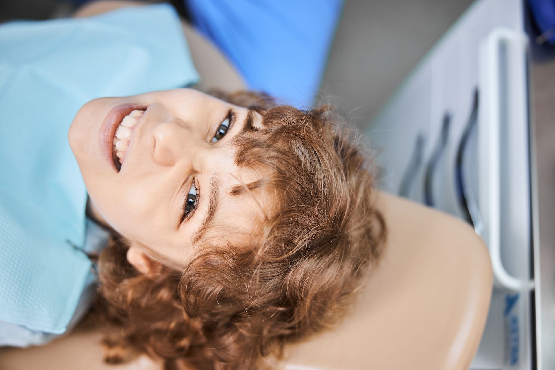 A person with curly hair smiles while lying back in a dentist's chair, wearing a light blue dental bib.