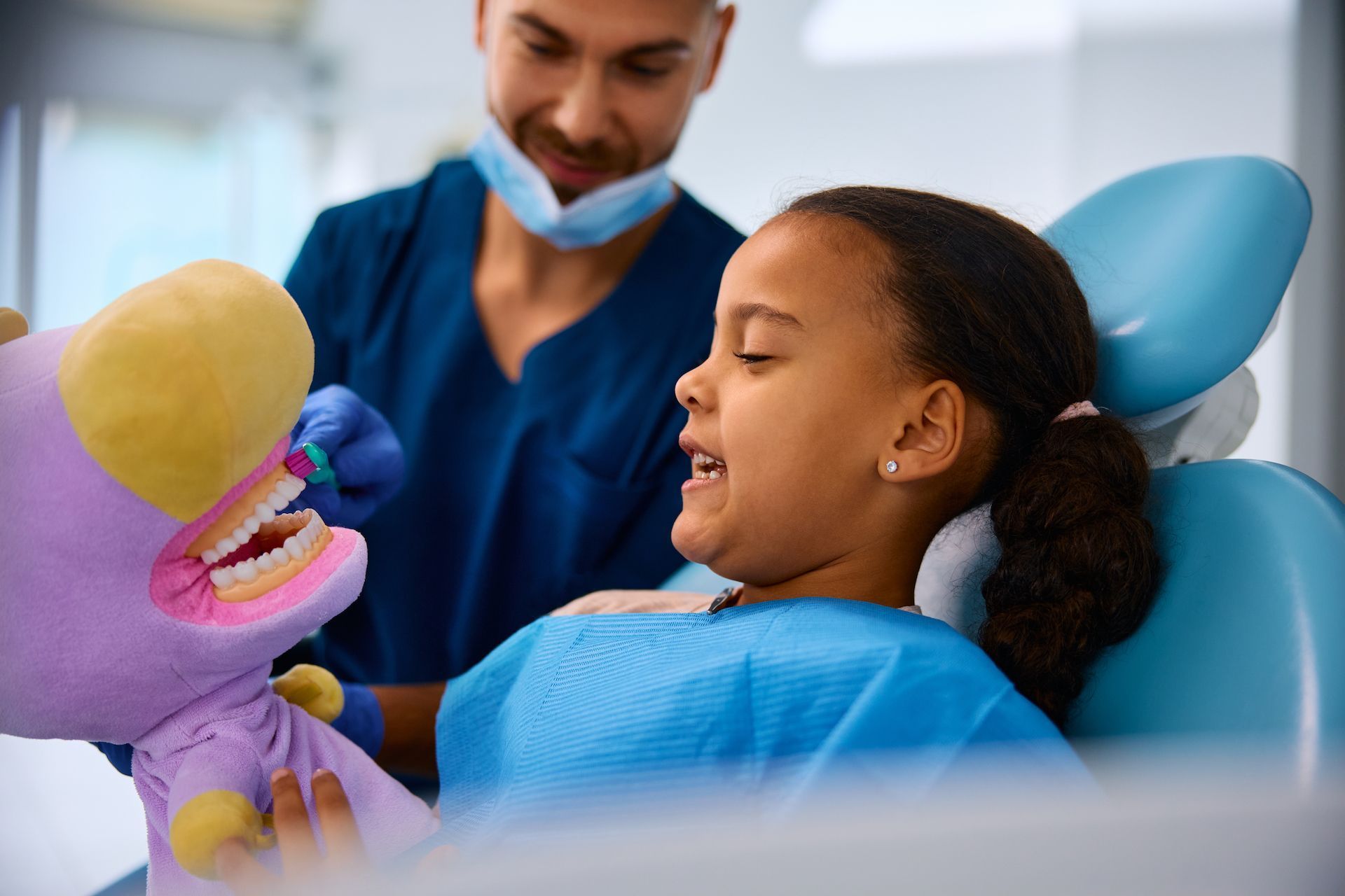 A dentist uses a plush puppet with human-like teeth to demonstrate proper brushing techniques to a child in a clinic.
