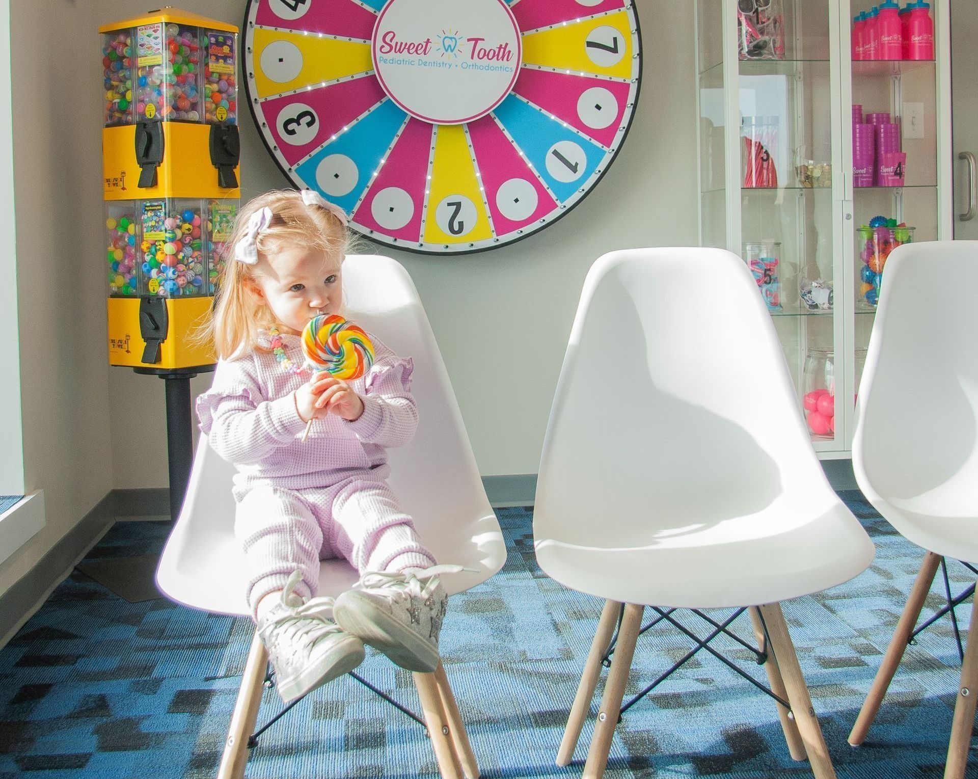 A toddler in a light purple outfit sits on a white chair, eating a colorful lollipop in a dental or play office.