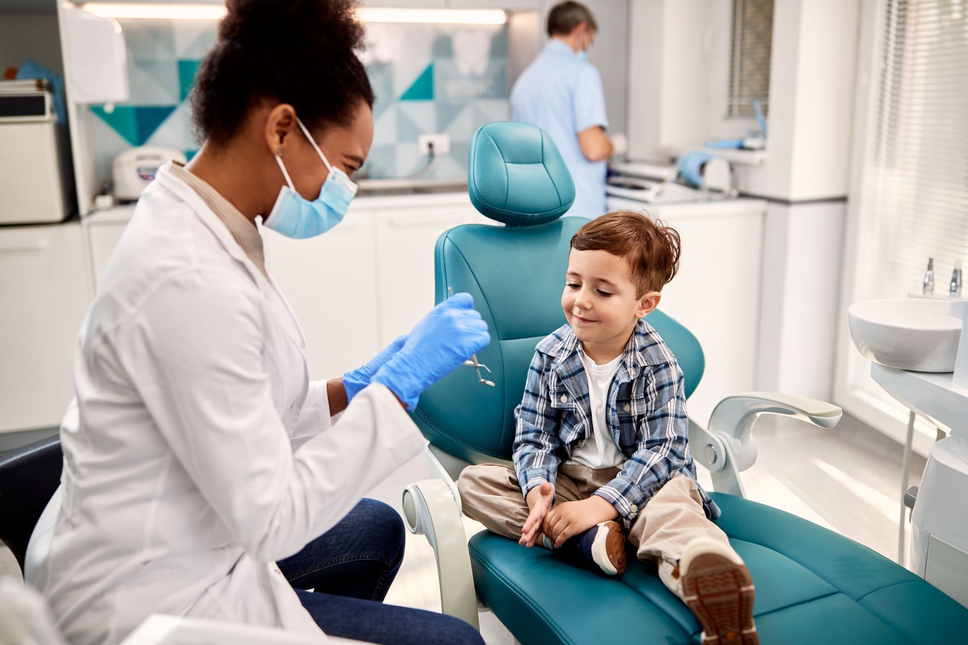 A dentist in a white coat and mask holds a tool while sitting with a smiling child in a dental chair.