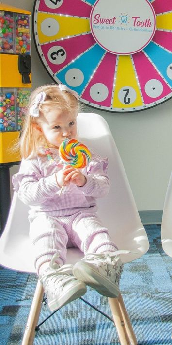 A toddler in a lavender sweater sits in a white chair, eating a colorful swirl lollipop in front of a prize wheel.