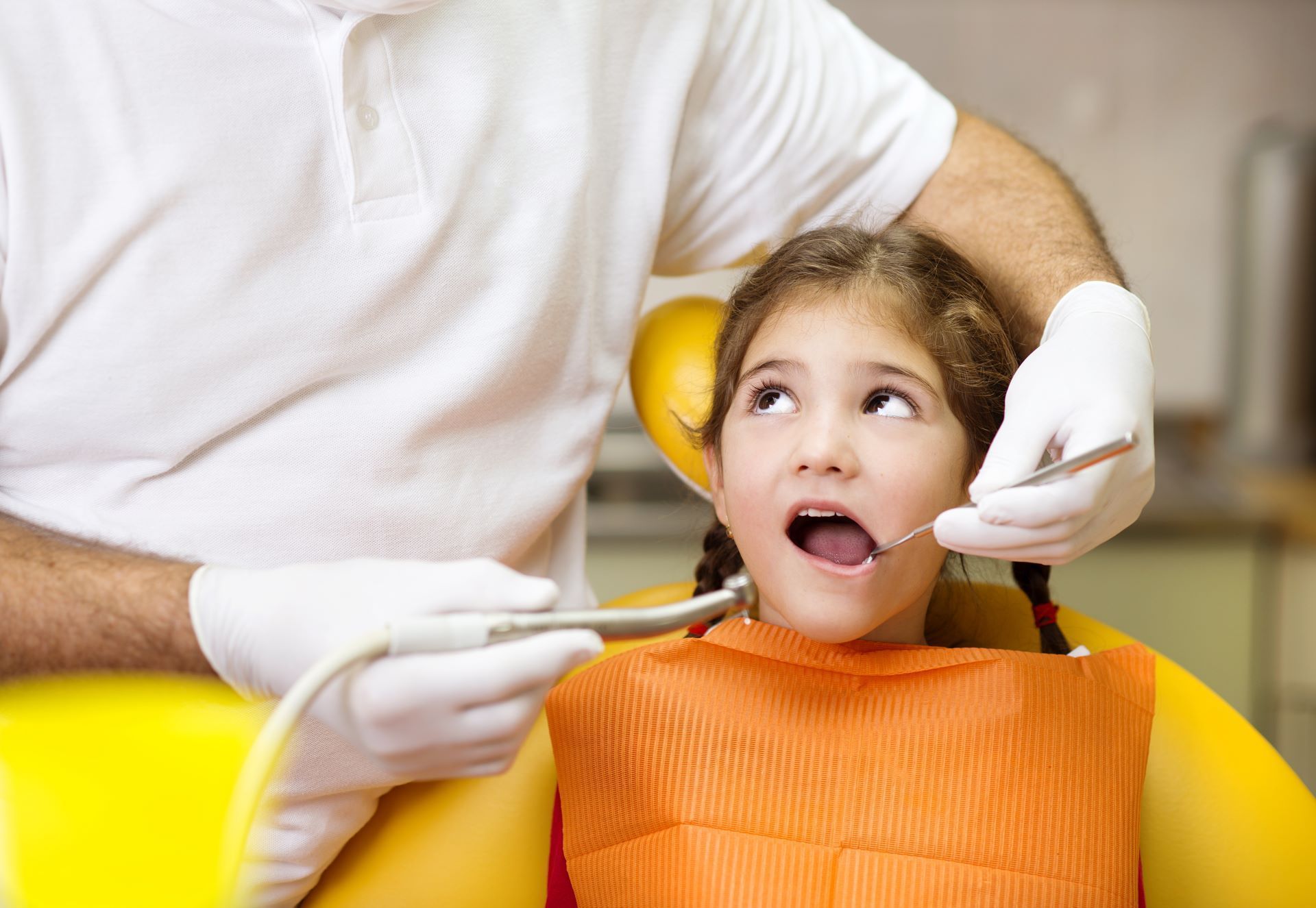 A dentist with white gloves examines a child in a yellow chair with an orange bib.