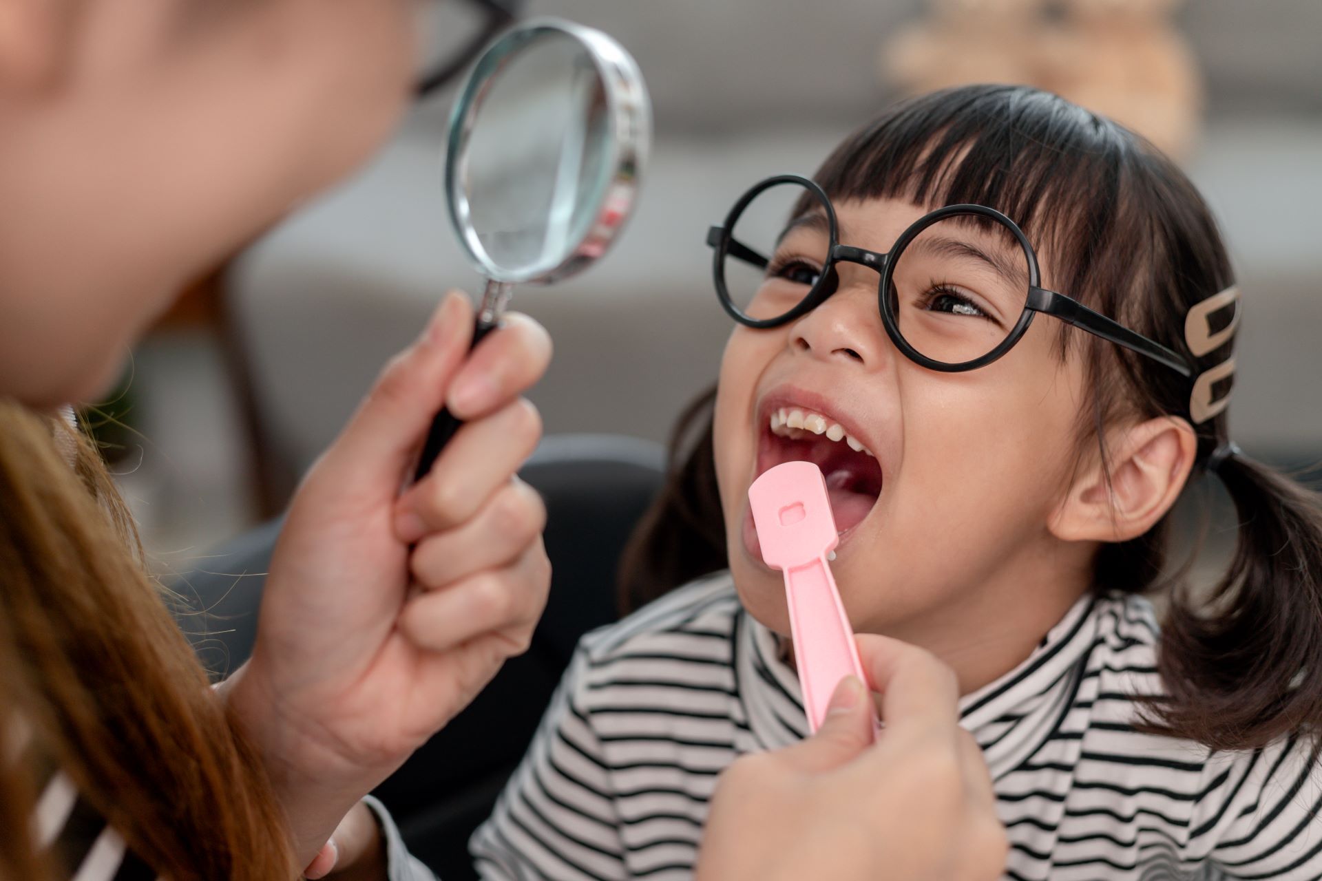 A person uses a magnifying glass and a pink tool to examine a smiling child wearing round glasses.