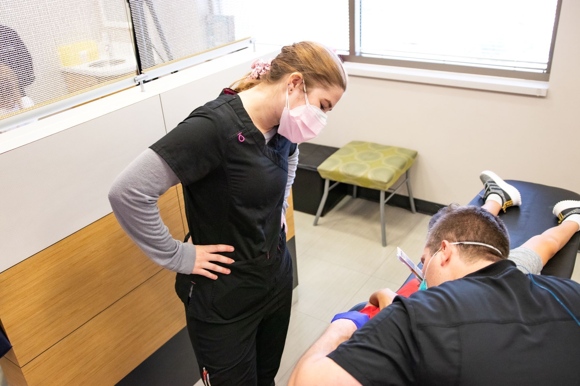 A medical professional in scrubs observes a patient lying on an exam table in a bright, clinical room.