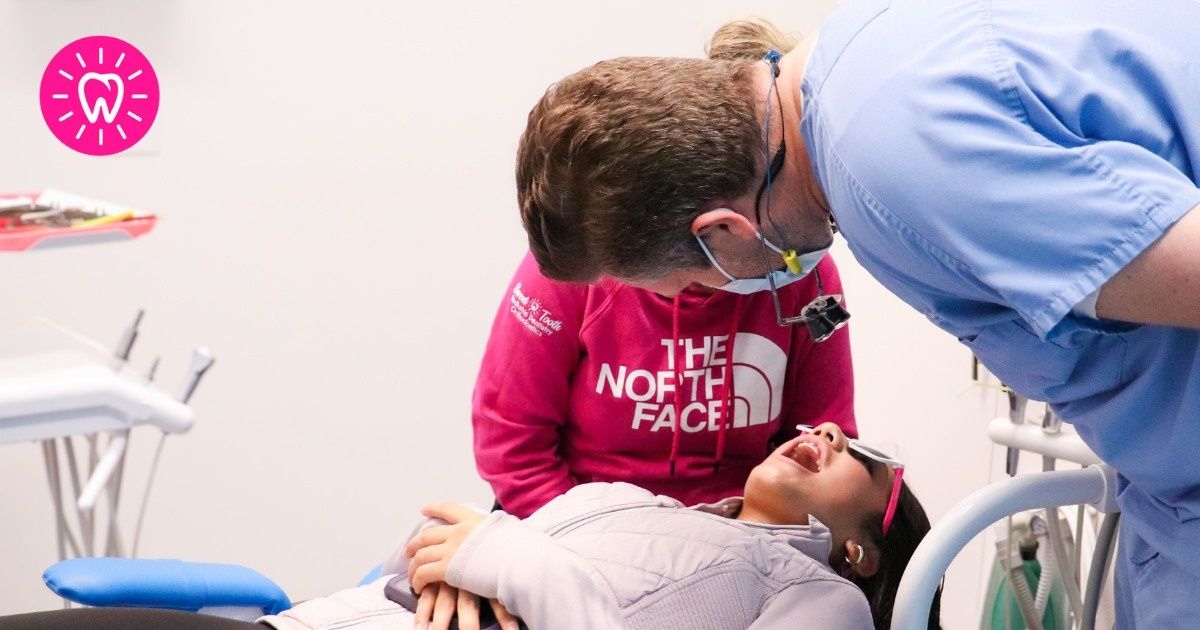 A dentist leans over a patient in a dental chair. The patient is looking up, mouth open, during a dental examination.