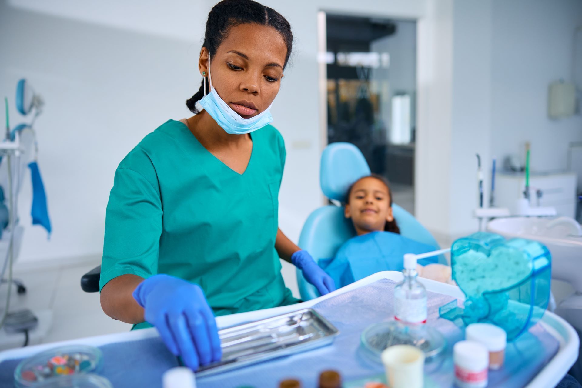 A dental professional in scrubs and a mask reaches for instruments on a tray, with a patient seated in the background.