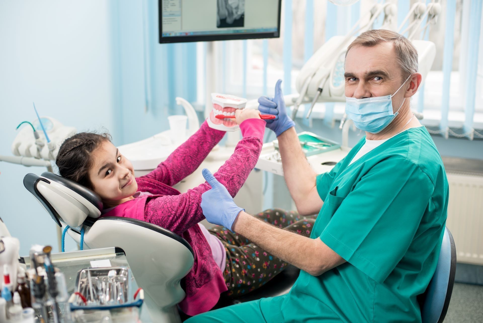 A smiling dentist and a young patient in a dental chair both give a thumbs up while holding a model of teeth.