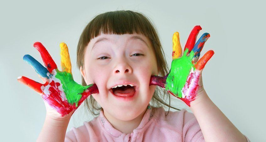 A child with Down syndrome smiling, holding up both hands painted with bright, colorful streaks of paint against their face.
