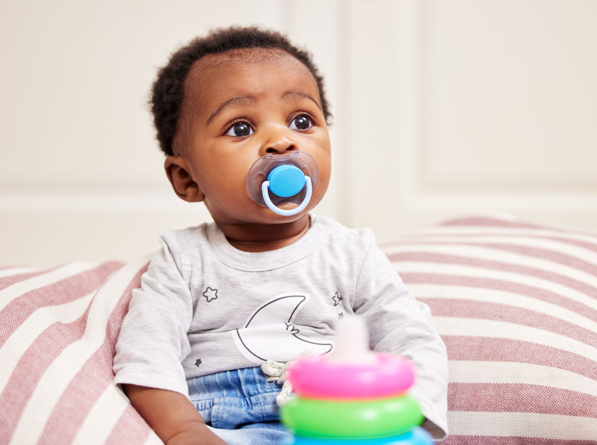 Baby with blue pacifier sitting on a pink-and-white striped cushion, looking off to the side.