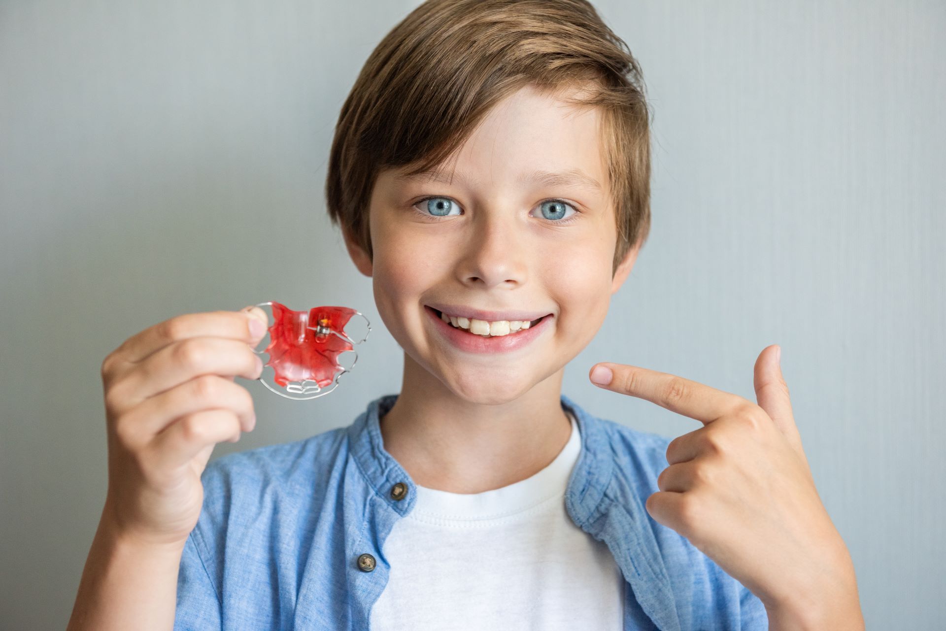 A child smiles while holding a red orthodontic retainer and pointing to their teeth.