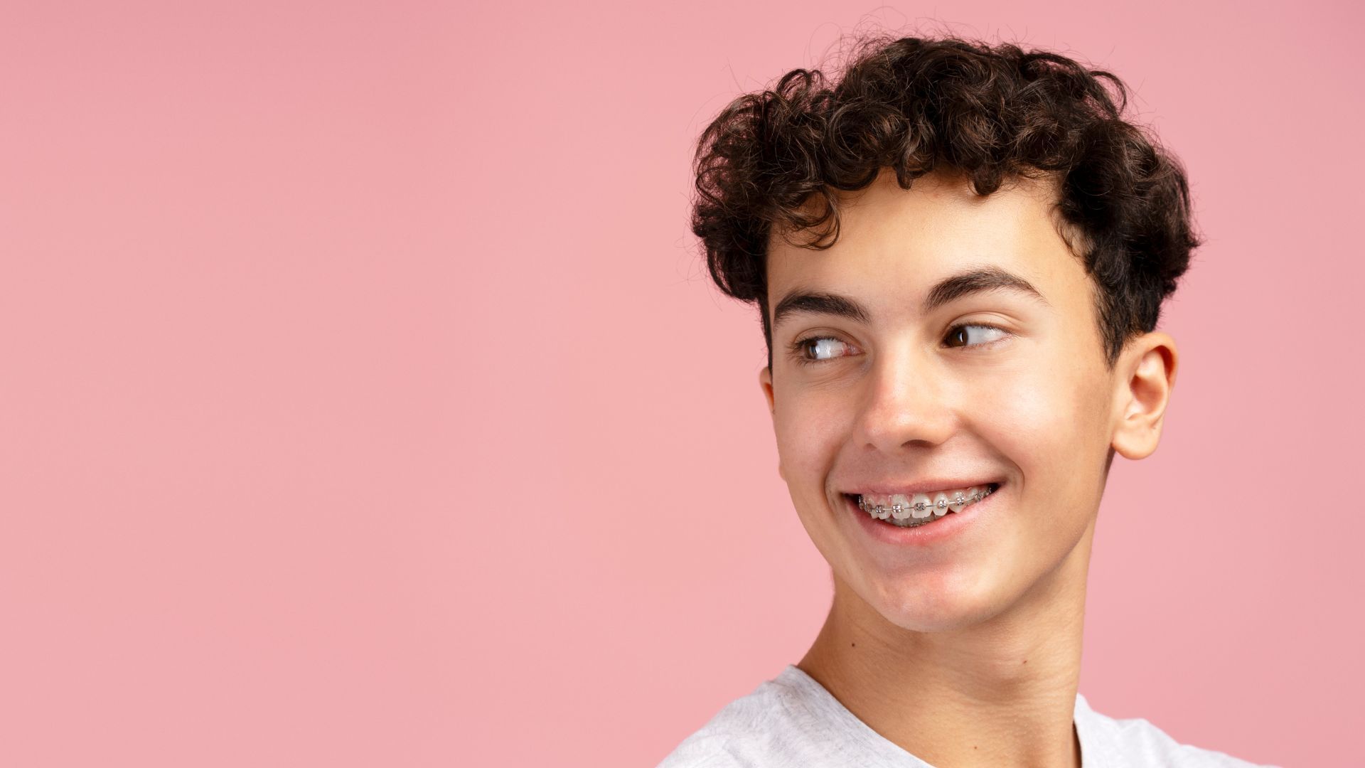 A young person wearing denim overalls and brown boots holding a lollipop in a dental office with hot pink walls.