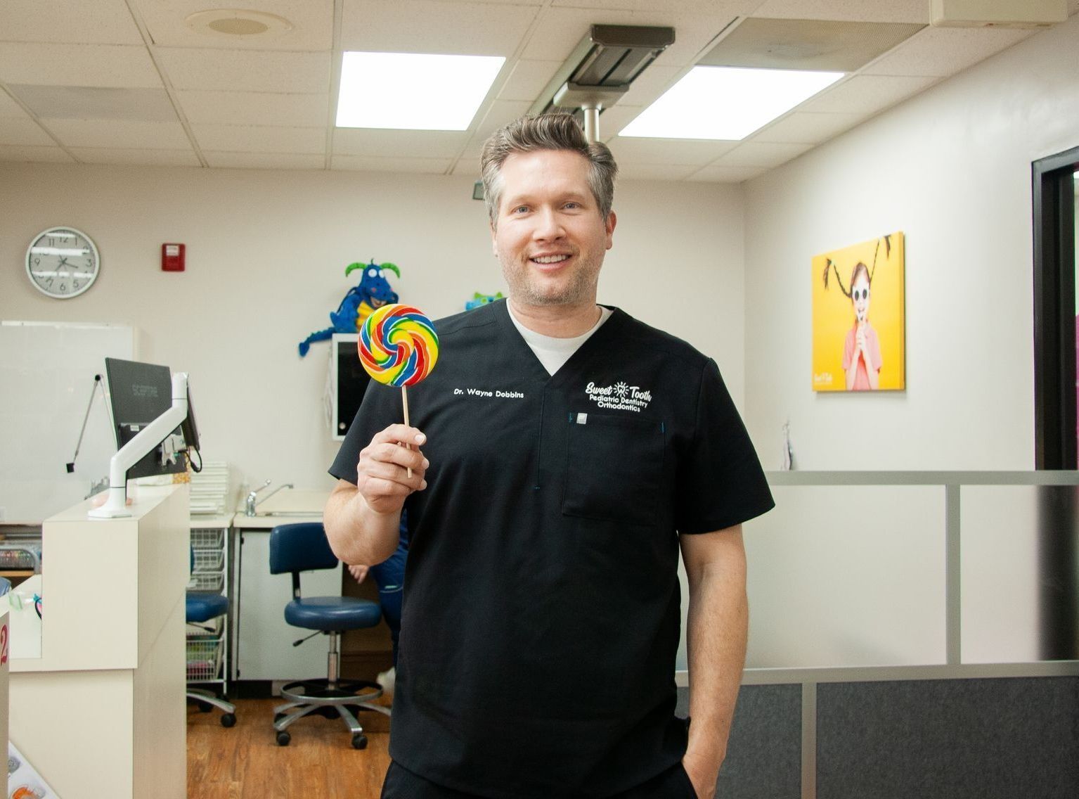 A person wearing black medical scrubs stands in a dental office holding a large, colorful lollipop.
