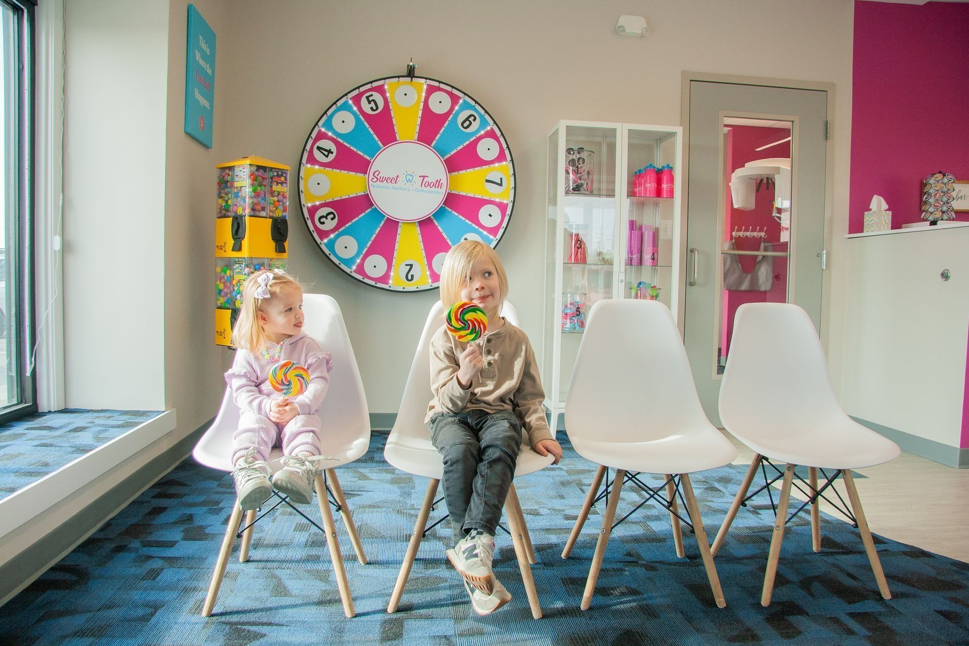 Two children sit on white chairs in a room with a colorful prize wheel, a toy dispenser, and a display cabinet.