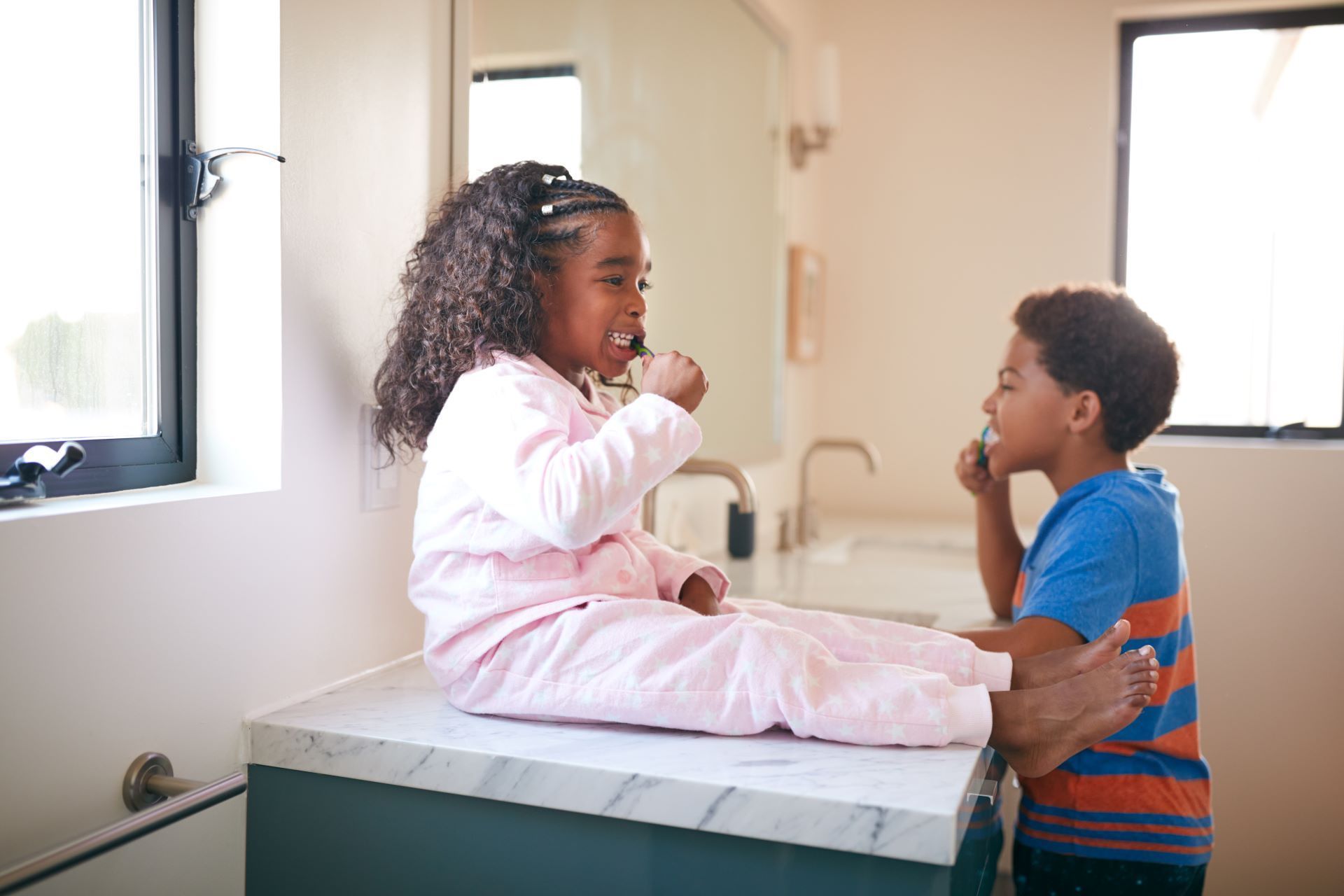 Two children brushing their teeth while sitting on a bathroom counter in a sunlit room.