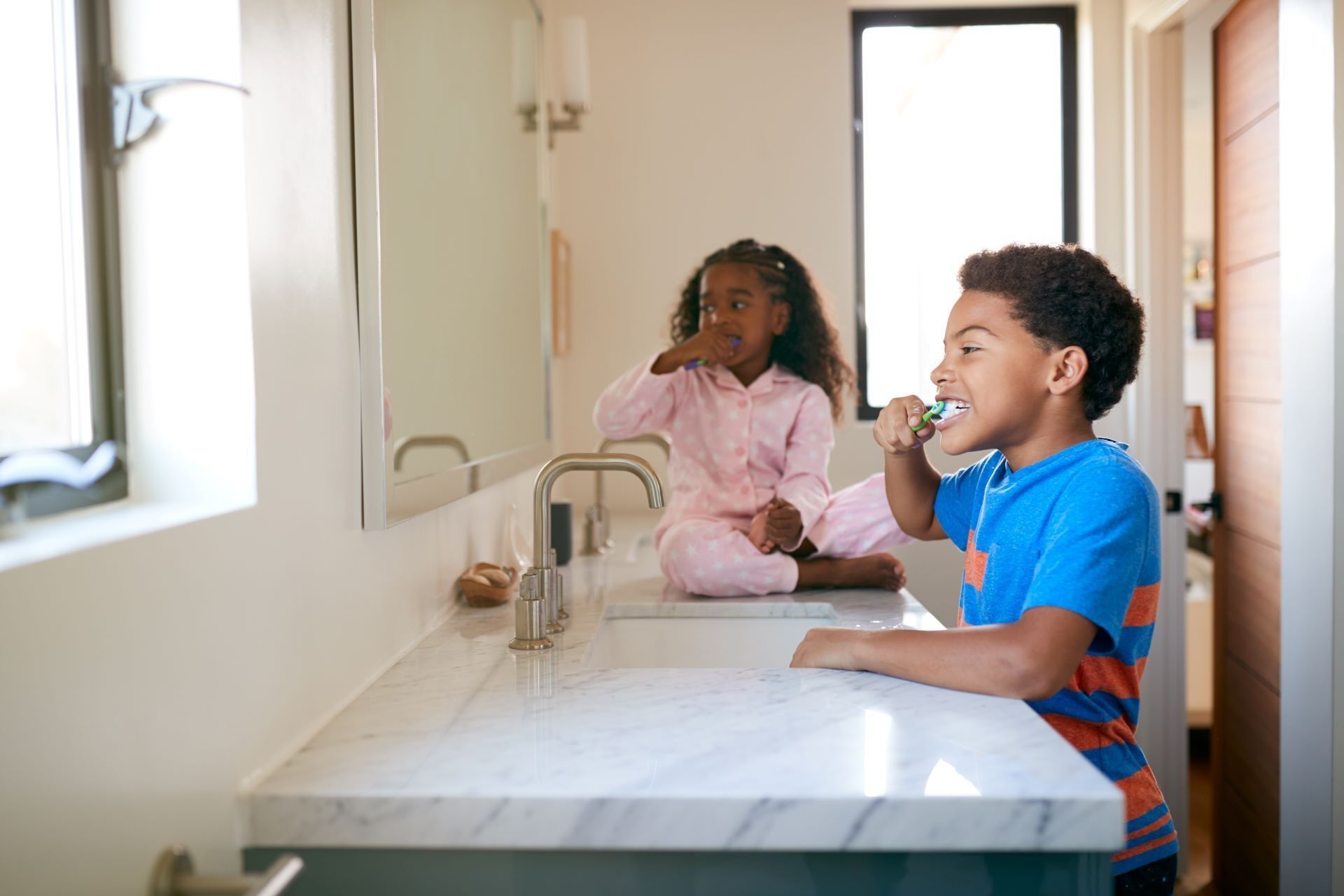 Two children in a bright bathroom, one brushing teeth while the other sits on the counter looking into a mirror.