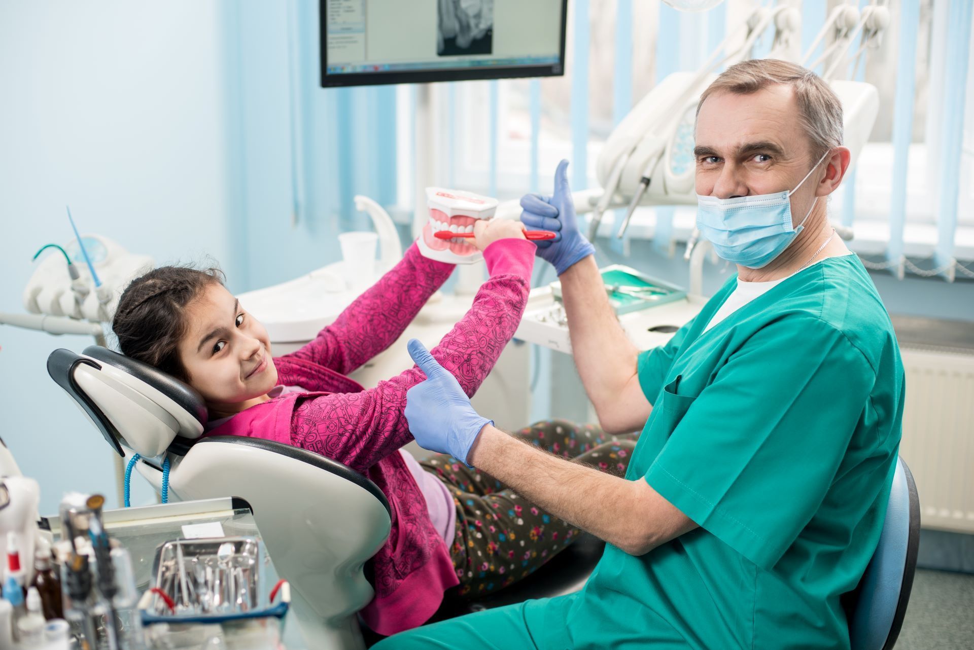 A smiling person in a dental chair and a dentist wearing a mask and scrubs give a thumbs-up while holding a model set of teeth.