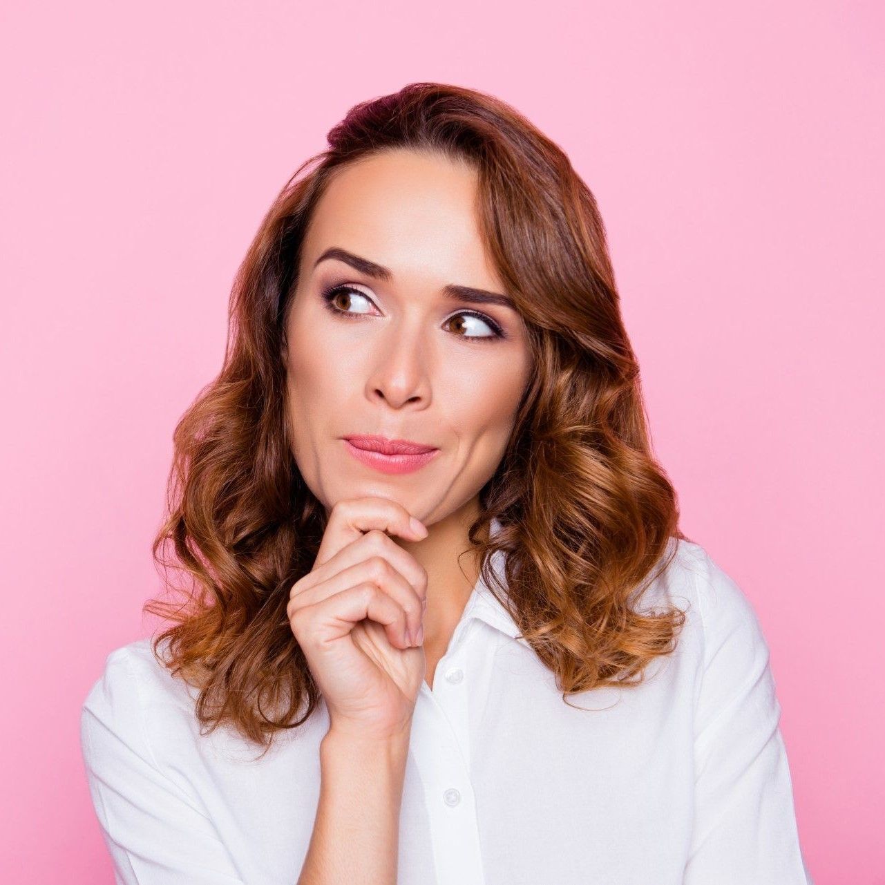 A person with shoulder-length wavy hair and a white shirt poses against a pink background with a thoughtful expression.