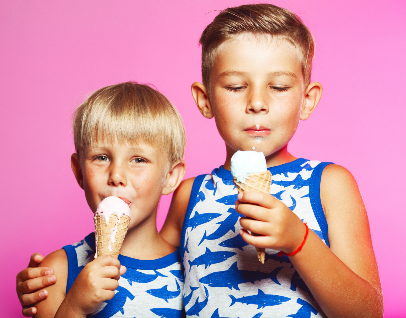 Two children in matching shark-print shirts eat ice cream cones against a solid pink background.