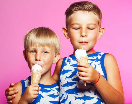 Two children in matching shark-print shirts eat ice cream cones against a solid pink background.