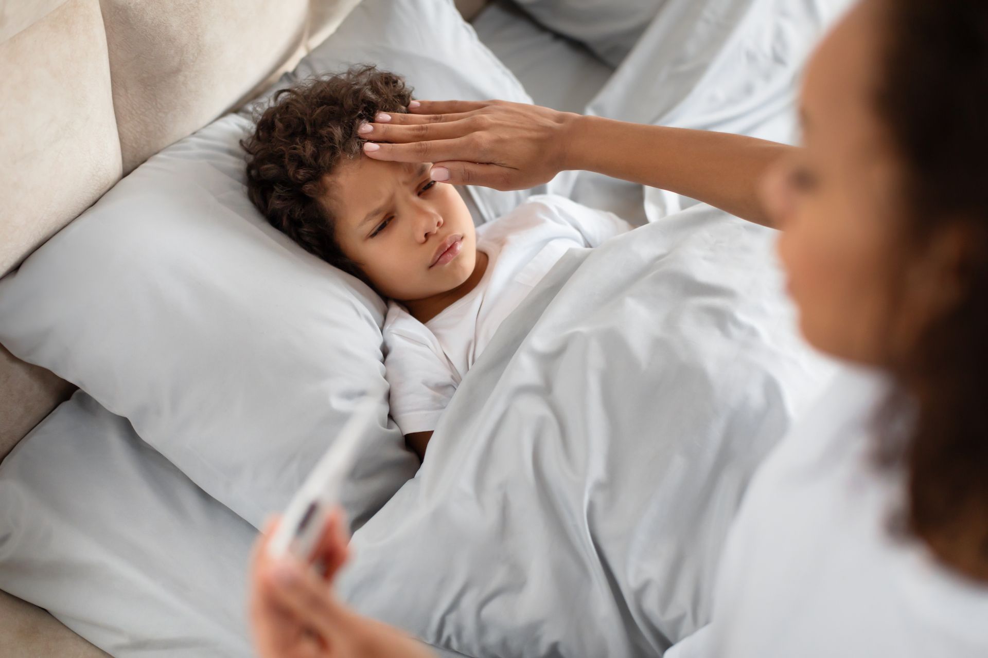 A person checking a child's forehead for fever while holding a thermometer in a bedroom.