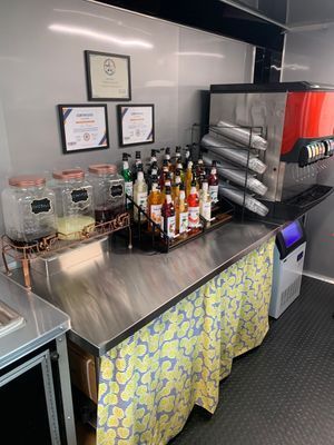 Mobile beverage station with syrups, juices, cups, and an ice machine, under a white wall, and a yellow skirt.