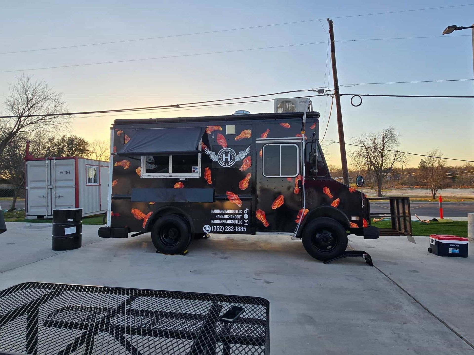 Food truck with black exterior and orange graphic parked outdoors, with a sunset in the background.
