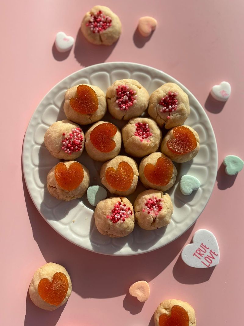 Plate of thumbprint cookies, some with red or orange hearts, on a pink surface with heart candies.