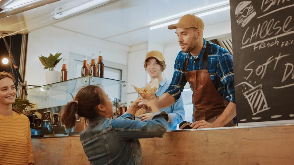 People at a food truck: a woman receiving a paper bag from a worker behind the counter.