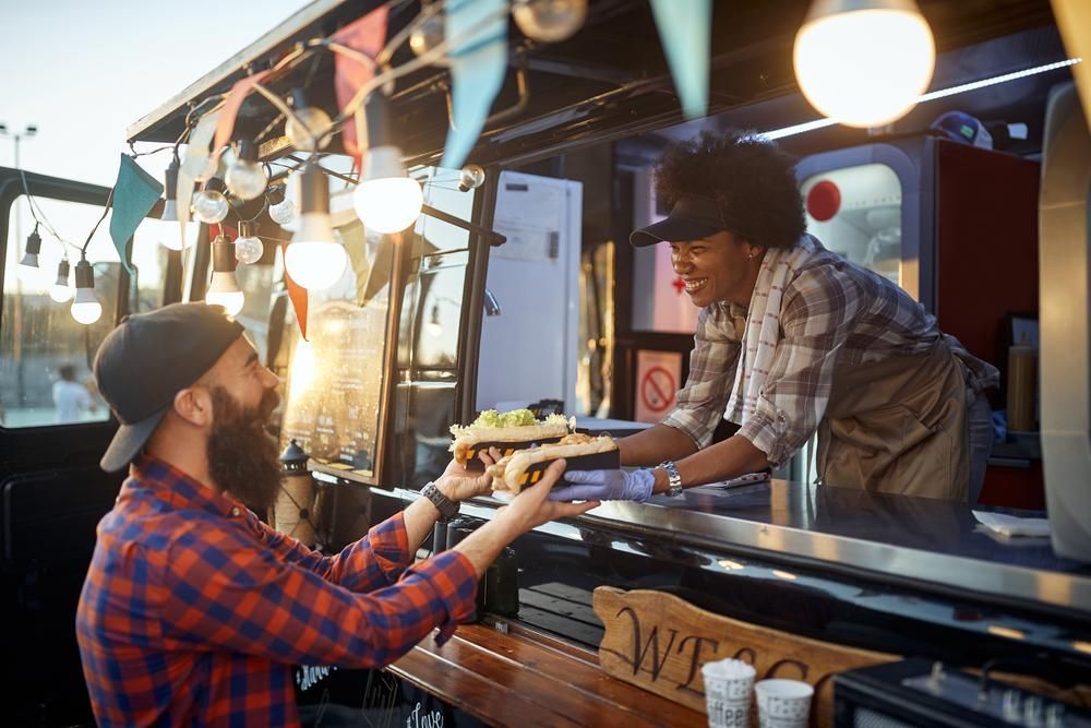 Person handing food to customer at food truck, warm lighting, outdoor setting.