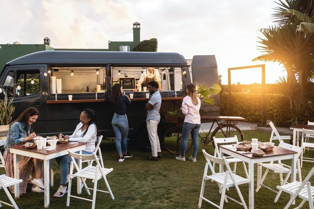 Food truck with customers, outdoor seating on a lawn. People order, eat, and socialize near a black van.