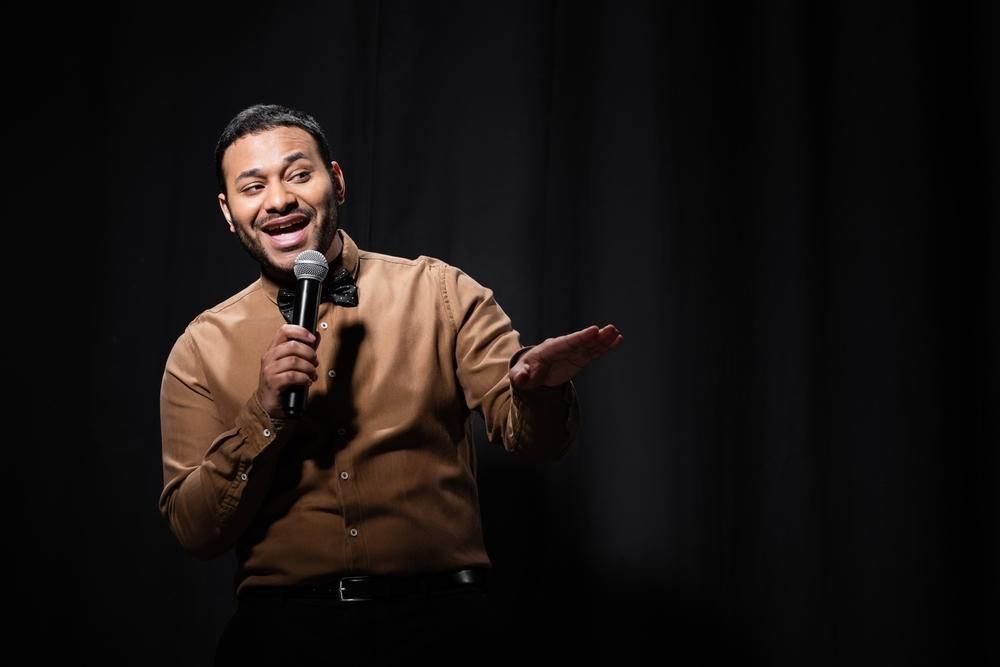Man with a microphone on stage, brown shirt, black bow tie, gesturing, smiling.